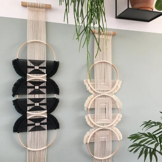 Deux décorations murales en macramé suspendues côte à côte sur un mur gris clair. Celle de gauche est principalement noire avec des motifs blancs, tandis que celle de droite est dans des tons écrus et beiges. Ces créations artisanales apportent une touche bohème et texturée à l'espace.