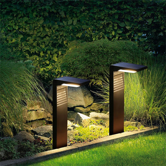 Deux lampes solaires extérieures Écolo illuminent un petit espace paysager composé de gazon, de plantes et de pierres. Leur design épuré et minimaliste projette une lumière chaleureuse et ambiante le long dun chemin le soir.