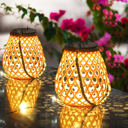 Deux lanternes Lampe solaire jardin haut de gamme-Bambou Tissé émettent une lumière jaune chaude sur une surface brillante, avec des bougainvilliers roses flous en arrière-plan, créant une ambiance extérieure chaleureuse et vibrante.