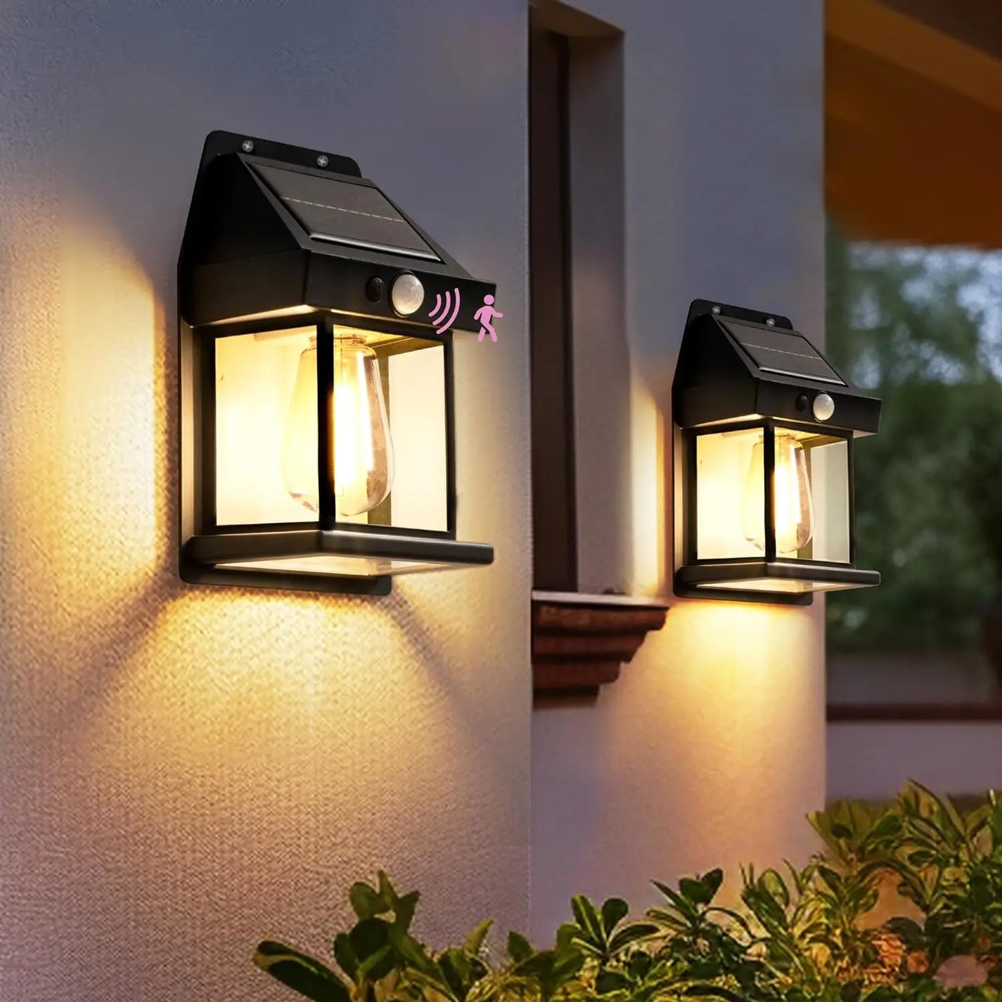 Deux luminaires Lampe solaire murale-Solar-LED™ avec détection de mouvement sont montés sur un mur extérieur, éclairant une zone de porche au-dessus de buissons verts au crépuscule.