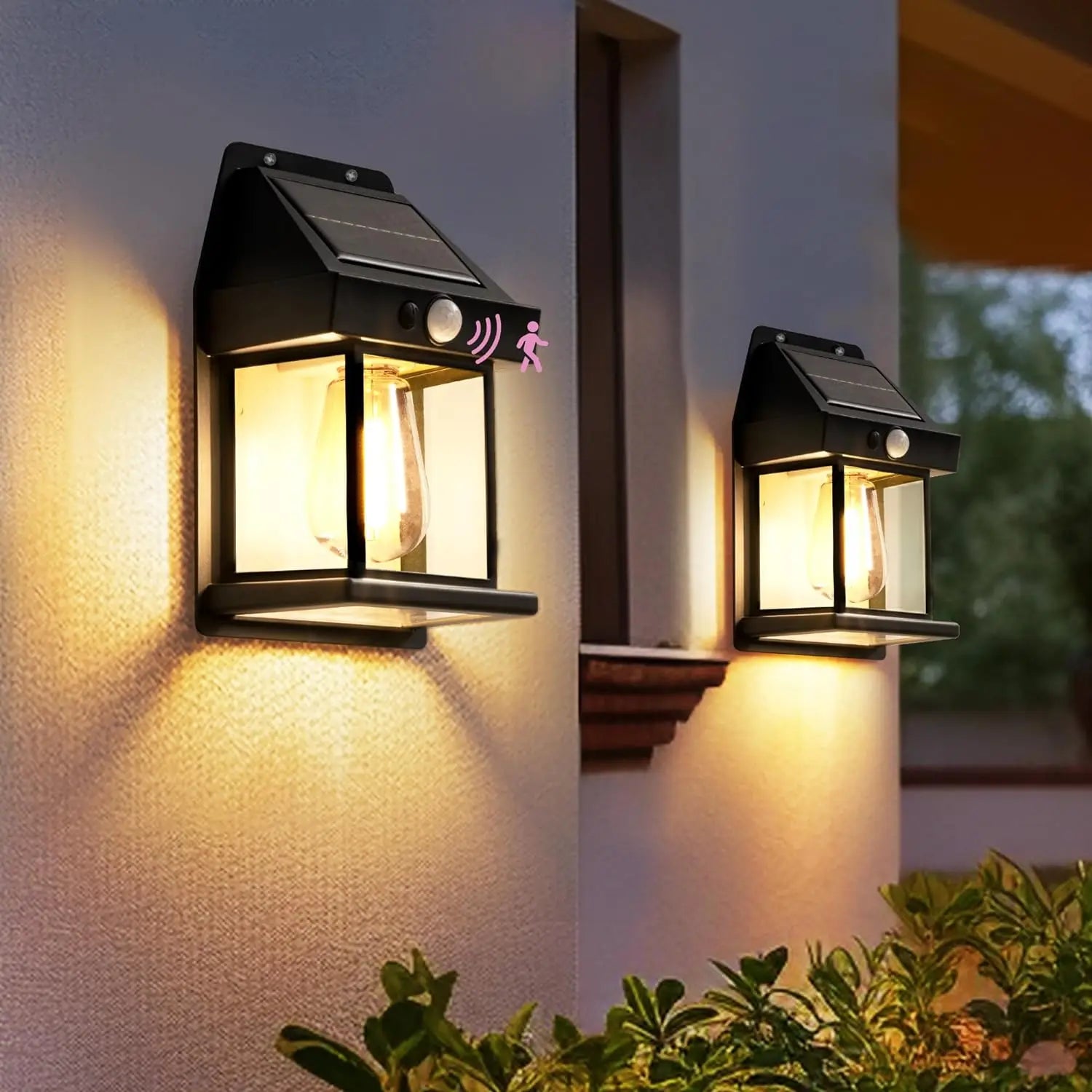 Deux luminaires Lampe solaire murale-Solar-LED™ avec détection de mouvement sont montés sur un mur extérieur, éclairant une zone de porche au-dessus de buissons verts au crépuscule.