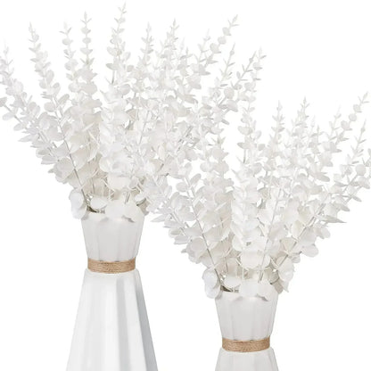 Deux vases en céramique blanche ornés de Plantes artificielles comme des vraies – Feuilles d'eucalyptus doré. Chaque vase présente un col étroit entouré de ficelle, idéal pour une décoration élégante sur un fond blanc uni.