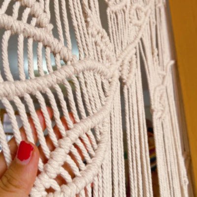 Une main avec un ongle rouge vif touche délicatement un  Macramé rideau  beige clair.
Les nœuds complexes et les motifs ajourés du rideau laissent entrevoir la lumière.
Le rideau, appelé "macramé rideau" en français, apporte une texture artisanale à l'espace.
Une décoration bohème et élégante capturée en détail.