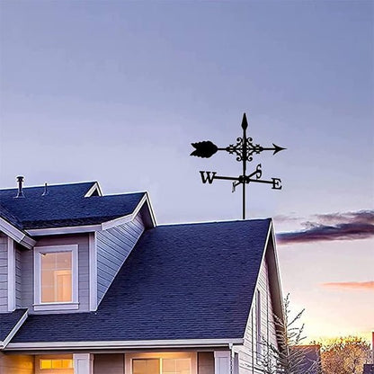 Une maison au toit bleu et aux garnitures blanches sous un ciel crépusculaire présente la Girouette de jardin-Galion, une girouette noire décorative au sommet, indiquant les directions cardinales avec une flèche et une silhouette de coq tandis que les nuages sont légèrement colorés par le soleil couchant ou levant.