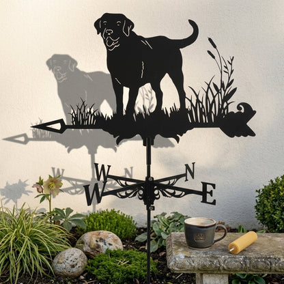 Girouette Animée en fer forgé noir représentant un Labrador, placée dans un jardin fleuri avec une maison en arrière-plan.