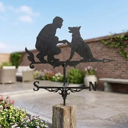 Girouette animal en fer forgé noir représentant un chien et son maître au-dessus des points cardinaux sous un ciel bleu.