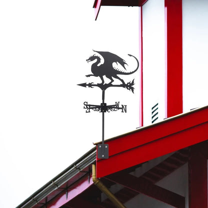 La Girouette-Dragon volant, une remarquable girouette noire en acier robuste, orne un bâtiment rouge et blanc. Cette décoration extérieure comprend des repères directionnels (N, E, S, O), avec le dragon perché sur une flèche, les ailes déployées, sur un ciel couvert.