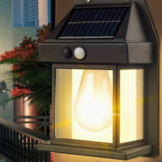 Gros plan sur la Lampe solaire murale-Solar-LED™, installée sur un bâtiment, avec son panneau solaire au sommet. Des plantes floues et un balcon en arrière-plan rehaussent l'ambiance élégante de cet éclairage extérieur.