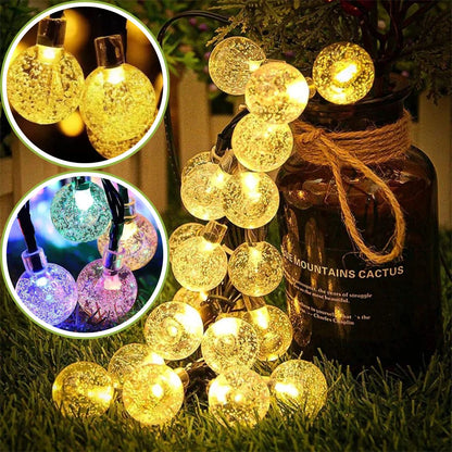 Les guirlandes lumineuses avec des ampoules rondes en verre craquelé brillent dans des tons chauds et froids. La guirlande solaire écologique | FlexiLumi est drapée sur un pot en verre brun étiqueté « Blue Mountains Cactus », illuminant une surface herbeuse. Les images encartées mettent en valeur les ampoules allumées, mettant en valeur leur fonction déconomie dénergie.
