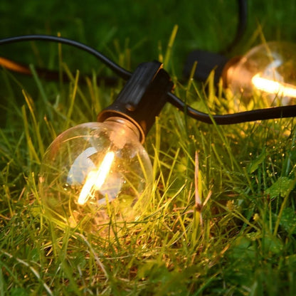 Une ampoule lumineuse sur une pelouse verte luxuriante, faisant partie de la Guirlande solaire extérieur puissante | Ambiance Enchantée. Deux ampoules visibles émettent une lumière chaude, renforçant lambiance magique, tandis que des cordons noirs les relient, contrastant avec lherbe vibrante.