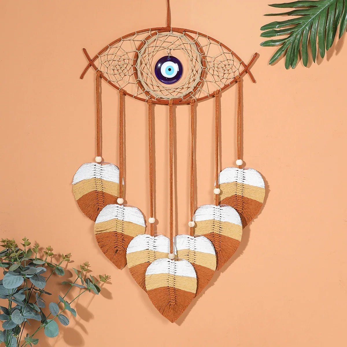 Un attrape-rêves décoratif en macramé en forme d'œil, orné d'une perle bleue en son centre, suspendu devant un mur pêche uni. De fines cordes en macramé couleur crème tissent une toile délicate à l'intérieur de l'anneau en bois clair. Cinq pendentifs en forme de feuille, réalisés en macramé dans des tons de crème, de blanc et de marron, pendent sous l'attrape-rêves, chacun terminé par une petite perle blanche.
