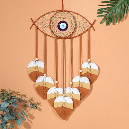 Un attrape-rêves décoratif en macramé en forme d'œil, orné d'une perle bleue en son centre, suspendu devant un mur pêche uni. De fines cordes en macramé couleur crème tissent une toile délicate à l'intérieur de l'anneau en bois clair. Cinq pendentifs en forme de feuille, réalisés en macramé dans des tons de crème, de blanc et de marron, pendent sous l'attrape-rêves, chacun terminé par une petite perle blanche.