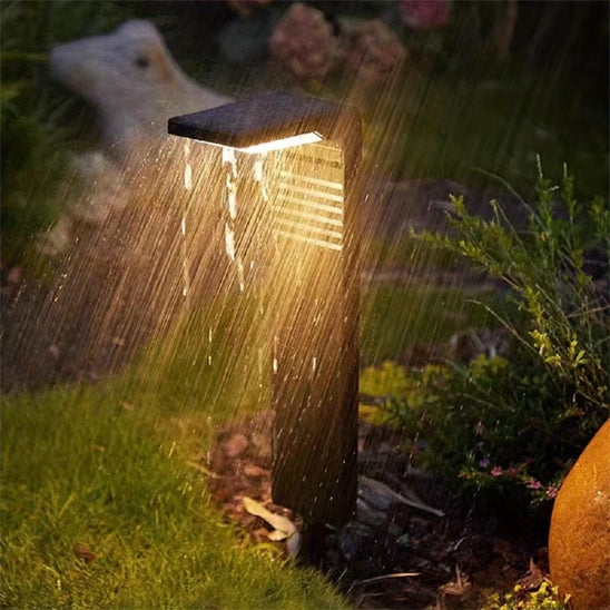 La Lampe solaire extérieur | Écolo illumine le jardin lors des averses, sa lumière chaleureuse mettant en valeur la verdure et une structure en pierre. A la fois économique et écologique, cet écrin serein prospère malgré les intempéries.
