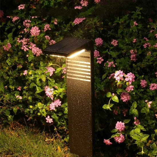 La Lampe solaire extérieure | Écolo illumine un parterre de fleurs roses et de feuillage vert grâce à son design moderne avec poteau rectangulaire incliné, diffusant une lueur chaleureuse et offrant une solution écologique et économique.