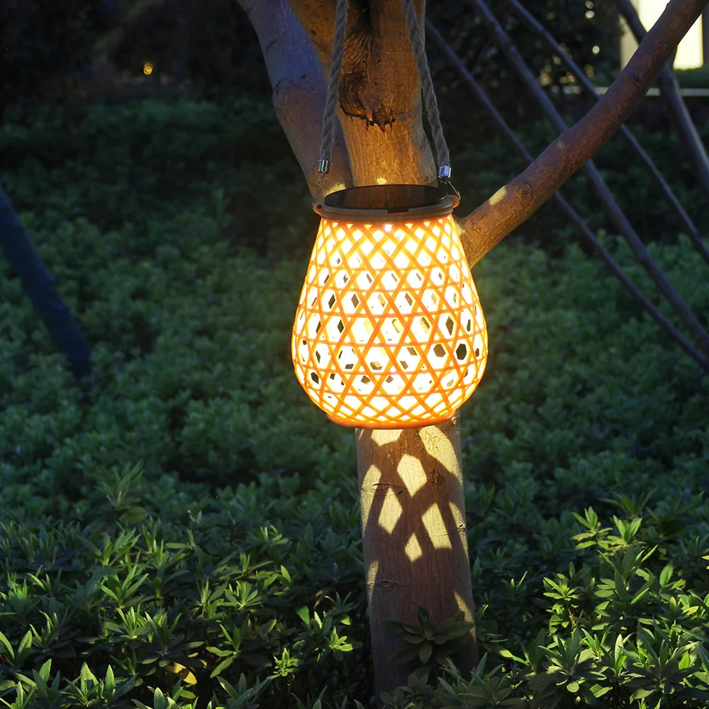 La Lampe solaire jardin haut de gamme-Bambou Tissé est suspendue à une branche d'arbre au crépuscule, projetant une lumière chaude et des motifs d'ombres complexes sur le tronc et le feuillage vert à proximité.
