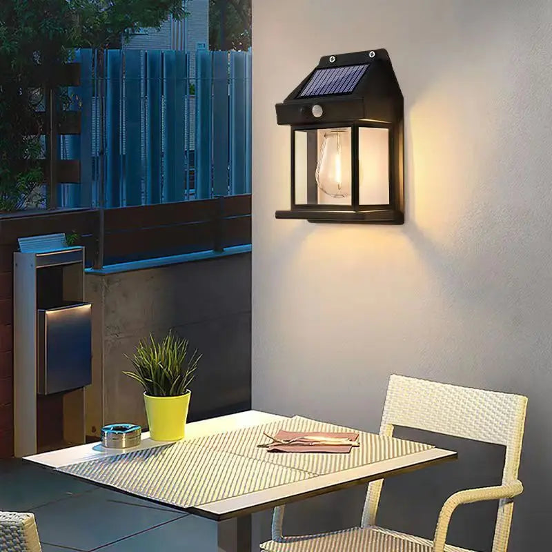 La Lampe solaire murale-Solar-LED™ éclaire une terrasse au crépuscule, illuminant une table avec plante en pot, livre et lunettes. Deux chaises blanches tressées complètent la scène devant une clôture extérieure.