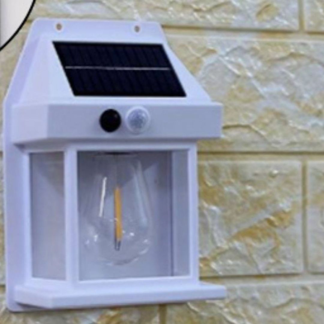 La Lampe solaire murale-Solar-LED™, dotée d'une ampoule en verre transparent et d'un petit capteur de mouvement noir, est montée sur un mur beige texturé en carrelage pour fournir un éclairage extérieur pratique.