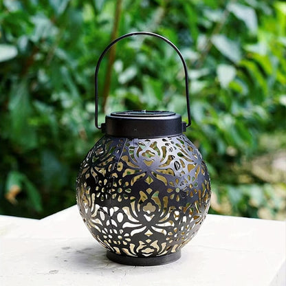 La lanterne solaire extérieure taupe de Mafrenesiedecoration est une lampe dextérieur décorative à énergie solaire avec des motifs découpés en métal complexes. Elle est dotée dune poignée supérieure et projette une lumière ambiante sur son environnement sur fond de feuillage vert luxuriant.