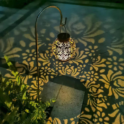 La lanterne solaire extérieure taupe de Mafrenesiedecoration projette des motifs floraux complexes sur une allée la nuit, entourée de fougères luxuriantes. Cette lampe élégante rehausse le charme et lambiance de votre jardin grâce à sa lumière solaire.