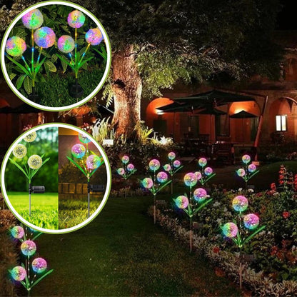 Une scène de jardin nocturne présente la Lampe solaire pissenlit | Ambiance LED de Mafrenesiedecoration, illuminant un chemin avec des lumières multicolores. Des gros plans révèlent des fleurs artificielles éclatantes mettant en valeur le jardin lumineux, tandis quun grand arbre et un bâtiment éclairé complètent la vue pittoresque.