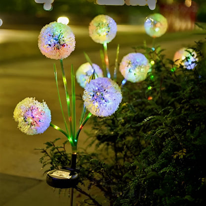 La Lampe solaire pissenlit | Ambiance LED de Mafrenesiedecoration illumine une allée de jardin dune lueur en forme de pissenlit, projetant des teintes de bleu, de vert, de rose et de jaune au milieu des plantes pour une ambiance déclairage écologique la nuit.