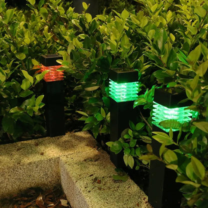 Des lumières de chemin de jardin au milieu dun feuillage vert luxuriant, brillant de couleurs rouges et vertes. Les lumières sont montées sur des poteaux noirs, illuminant le bord dun chemin en pierre la nuit. La scène est sereine et accueillante.