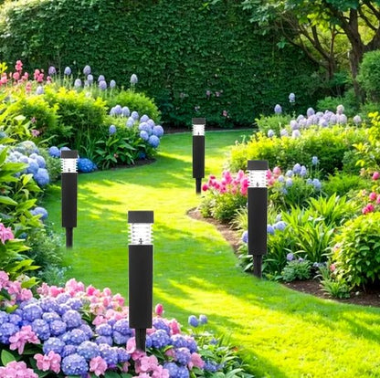 Un jardin luxuriant avec un chemin sinueux bordé de fleurs colorées, dont des hortensias roses et bleus. Plusieurs lampes solaires noires sont régulièrement espacées le long du chemin, avec des arbustes et des arbres verts en arrière-plan.
