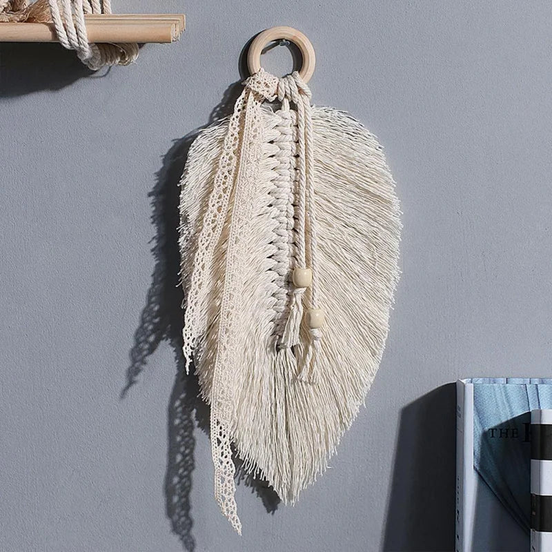 Le Macramé-Feuille est une tenture murale décorative avec deux feuilles de coton beige, un ruban de dentelle et des perles en bois, présentée sur un mur gris au-dessus d'une étagère.