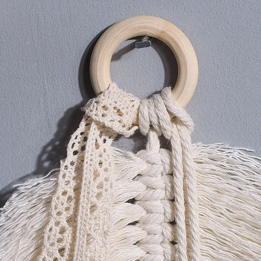 Un gros plan du Macramé-Feuille montre un anneau en bois avec des cordons et de la dentelle blanc cassé noués, avec des accents de feuilles de coton, formant une décoration murale texturée sur un fond gris clair.