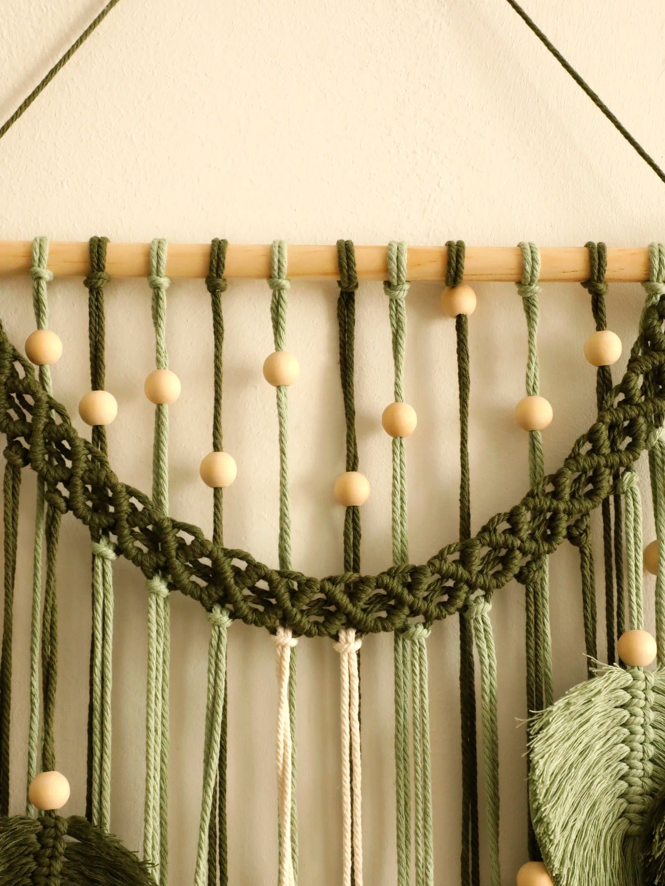 Macramé décoratif suspendu à un mur blanc, fait de cordes vert olive, vert clair et crème, orné de perles de bois clair. La texture nouée complexe du macramé crée un motif bohème. "Marcrame" apparaît subtilement intégré dans le design.