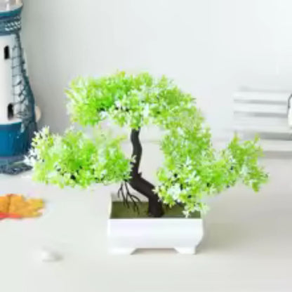 Sur cette image, on observe une plante artificielle de type bonsaï. Ses feuilles vertes et claires sont disposées de manière ordonnée. Elle est placée dans un pot carré blanc. À l'arrière-plan, on distingue d'autres éléments décoratifs sur une surface claire. Cette plante artificielle apporte une touche de verdure sans nécessiter d'entretien.