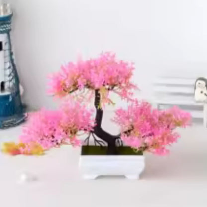 Sur cette image, on observe une plante artificielle ressemblant à un bonsaï en fleurs roses. Elle est placée dans un pot carré blanc. En arrière-plan flou, on distingue un phare miniature bleu et blanc, ainsi que d'autres objets décoratifs sur une surface claire. La lumière ambiante met en valeur les couleurs vives de la plante. Cette composition suggère un arrangement décoratif intérieur.