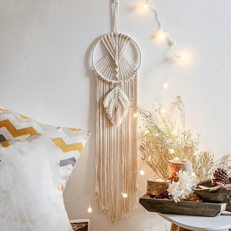Un attrape-rêves en macramé ivoire orne un mur blanc, apportant une touche bohème.
Des motifs complexes en cordes forment un cercle et de longues franges décoratives.
Une guirlande lumineuse douce et des éléments de décoration naturels complètent l'ambiance chaleureuse.
Macramé décoratif, une pièce artisanale élégante pour la maison.