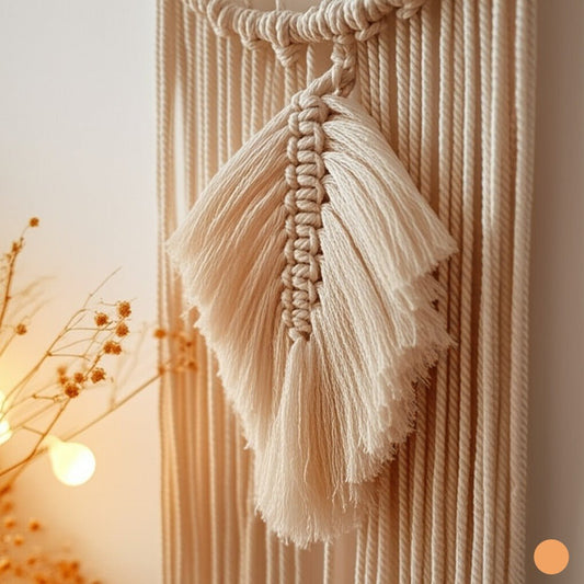Gros plan d'une décoration murale en macramé beige clair, présentant des nœuds complexes formant un motif texturé et une grande plume centrale détaillée.
Les longues franges de fils pendants ajoutent une dimension tactile et aérée à la pièce.
L'artisanat délicat met en valeur la beauté des fibres naturelles et le savoir-faire du nouage décoratif.
Une branche de fleurs séchées floue et une douce lumière en arrière-plan suggèrent un intérieur chaleureux et artistique.