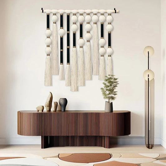 Rideau en macramé texturé ivoire suspendu à un bâton en bois clair, créant un point focal bohème sur un mur blanc.
Les détails noués complexes présentent des motifs géométriques et de longues franges douces.
Une commode en bois à lattes se trouve en dessous, ornée de petites sculptures et d'une plante en pot.
Un lampadaire moderne aux formes circulaires se dresse à droite, éclairant doucement la scène minimaliste et élégante.