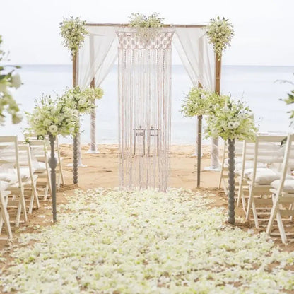 Un élégant décor de mariage sur une plage de sable clair, encadré par une arche en bambou drapée de tissu blanc vaporeux. Au centre, un délicat macramé rideau crème flotte doucement, créant un point focal romantique. Des bouquets luxuriants de fleurs blanches ornent l'arche et des arrangements floraux plus importants se dressent sur des piédestaux gris le long de l'allée, parsemée de pétales blancs.