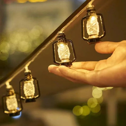 Une main se tend vers la GUIRLANDE EXTERIUR SOLAIRE de Lumière Heureuse, avec ses petites lanternes lumineuses qui rappellent doucement les décorations de Noël. Le fond est agrémenté de lumières floues, créant une atmosphère chaleureuse et ambiante.