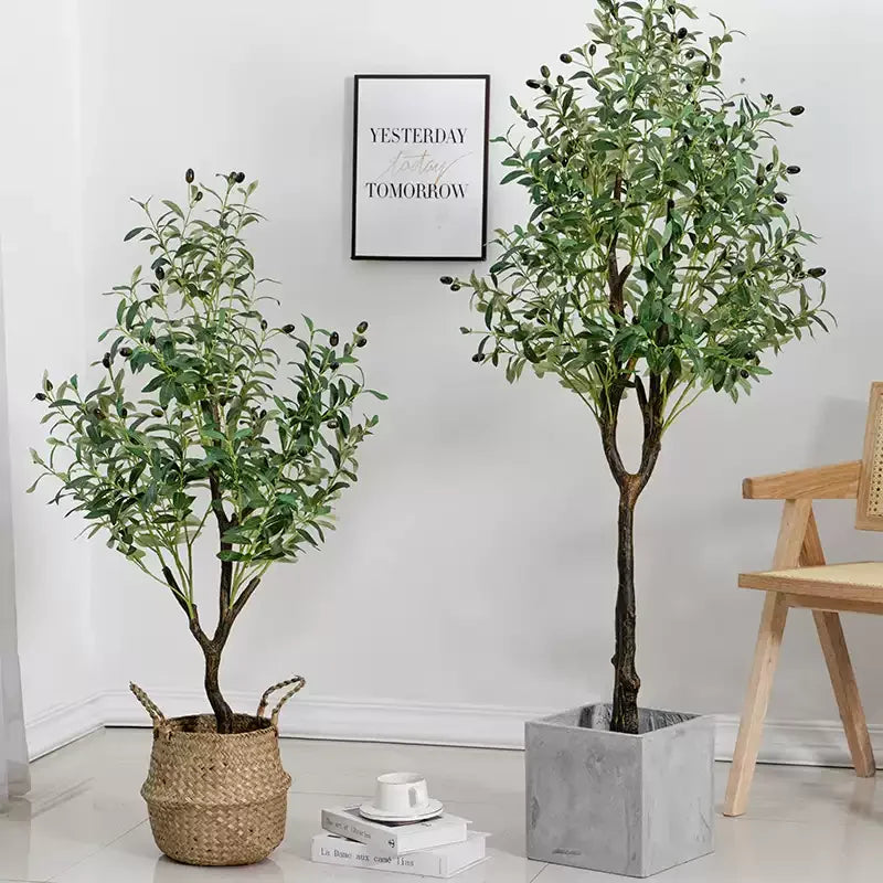 Deux arbres d'intérieur en pot, dont la Plante artificielle d'extérieur-Olive, sont posés sur un sol blanc près d'une chaise en bois. L'un est dans un panier tressé, l'autre dans un pot gris. Une décoration chaleureuse et une citation encadrée créent une ambiance de jardin intérieur accueillante.