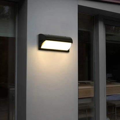 Lapplique murale « inégale LED » de Mafrenesiedecoration projette une lueur chaleureuse sur un mur gris près dune grande fenêtre et dune porte vitrée, illuminant lextérieur du bâtiment avec style et efficacité énergétique. Le décor semble être celui du soir ou du début de la nuit.