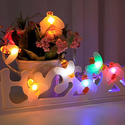 Abeilles Solaires : Des papillons et des champignons lumineux LED colorés illuminent une petite plante en pot aux fleurs roses sur un support blanc décoratif. Alimentées par l'énergie solaire, elles éclairent doucement l'arrière-plan, mettant en valeur des teintes vibrantes de manière écologique.