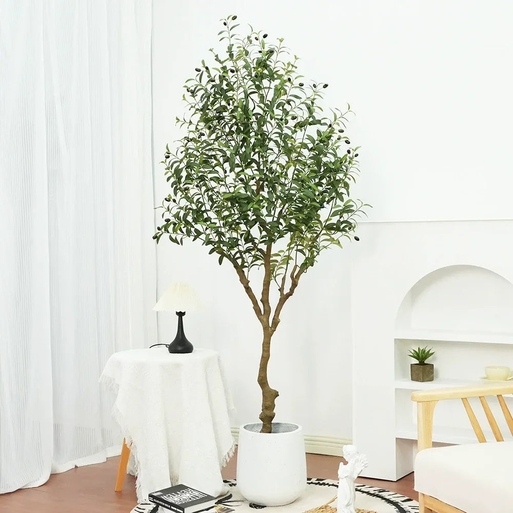 Une plante artificielle d'extérieur - Olive aux feuilles vertes se dresse sur un tapis à motifs dans une pièce lumineuse, à côté d'une table ronde avec une lampe, d'une petite plante sur une étagère et d'une chaise en bois.