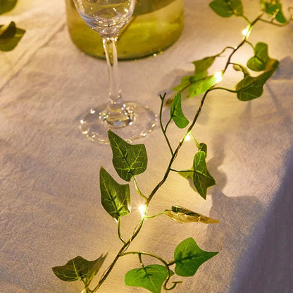 La Guirlande solaire extérieur | Lueur de bonheur, composée de lumières LED chaudes en forme de lierre entrelacées de feuilles vertes, orne une nappe blanche. Un verre à vin et une bouteille verte en arrière-plan complètent la scène chaleureuse et élégante.