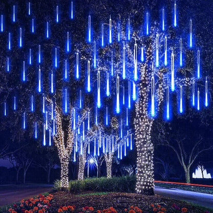 Des arbres ornés de lumières LED bleues en cascade et de troncs blancs scintillants créent une atmosphère magique et festive. Des fleurs multicolores s'épanouissent à la base tandis que la guirlande solaire extérieur Joie Solaire projette une lueur écologique sous le ciel nocturne sombre.