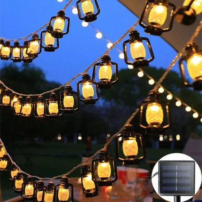 La GUIRLANDE EXTERIUR SOLAIRE | Lumière Heureuse est composée de petites guirlandes lumineuses en forme de lanternes qui illuminent chaleureusement un décor extérieur. Elégamment suspendues, les arbres et le ciel dégagé renforcent son charme festif. Un insert met en valeur un panneau solaire LED, probablement à l'origine de ces lumières enchanteresses.