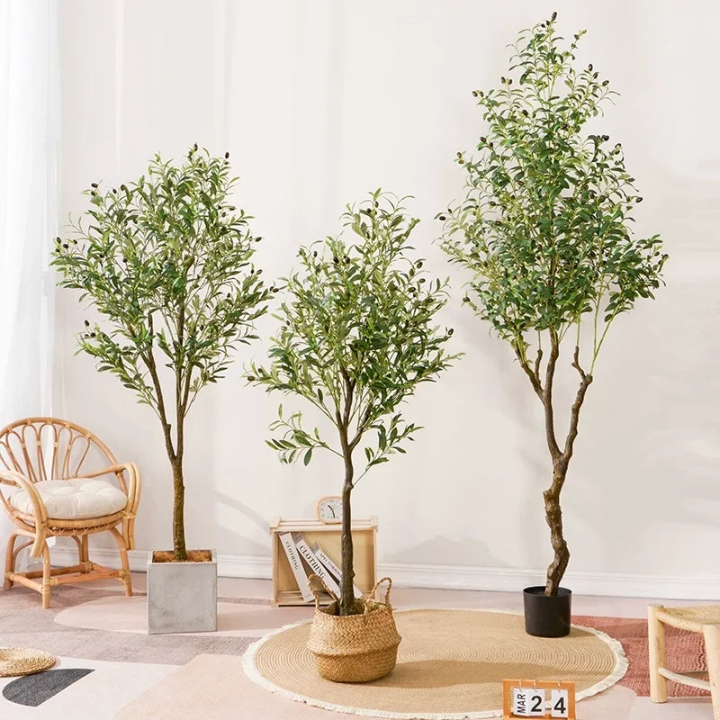 Trois Plantes artificielles d'extérieur - Oliviers de différentes hauteurs sont placés à l'intérieur sur un tapis léger, avec une chaise en osier et un décor neutre en arrière-plan.