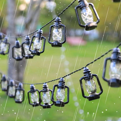 La GUIRLANDE EXTERIUR SOLAIRE de Lumière Heureuse, en forme de petites lanternes, se suspend élégamment à l'extérieur sous la pluie. Avec un fond vert flou, ces charmantes lumières ressemblent à des décorations de Noël tandis que les gouttes de pluie tombent doucement autour d'elles.