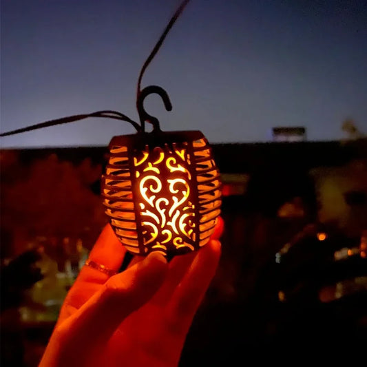 Une main tient une guirlande extérieure solaire | Flamme avec un effet flamme qui brille sur un ciel sombre. Ses volutes décoratives pendent d'un crochet, avec l'arrière-plan flou soulignant l'ambiance extérieure enchanteresse.