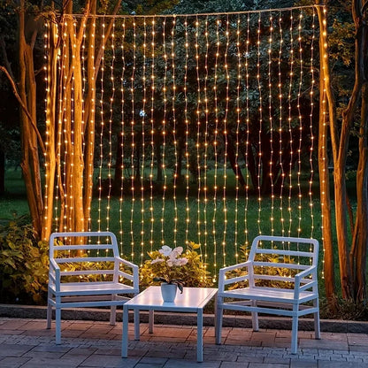 Scène de patio extérieur avec deux chaises blanches et une petite table. Des guirlandes lumineuses chaudes sont suspendues verticalement aux branches des arbres, créant une toile de fond lumineuse. La verdure luxuriante évoque une atmosphère chaleureuse et accueillante.