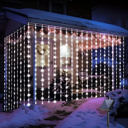 Un porche orné de guirlandes lumineuses en forme de glaçons et d'une guirlande solaire extérieur-rideau | Lumière heureuse crée une scène festive et hivernale. La neige recouvre le sol et les buissons, tandis qu'une couronne orne la porte dans cette présentation enchanteresse.