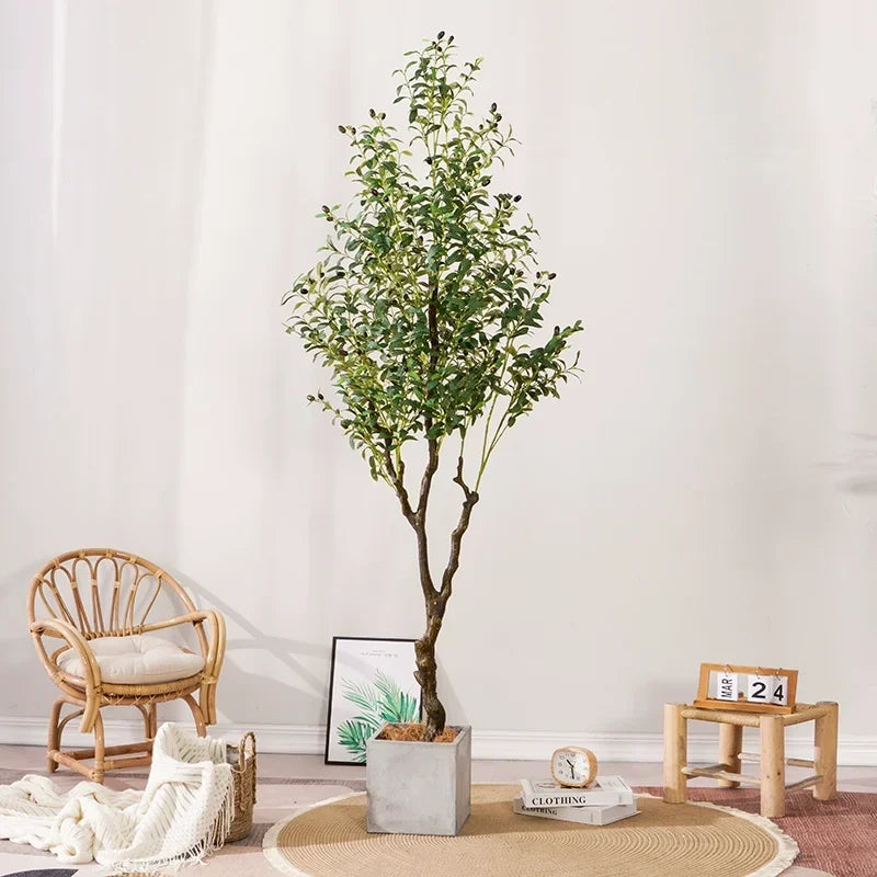 Une plante artificielle d'extérieur - Olive se dresse sur un tapis circulaire à côté d'une chaise en rotin avec un coussin blanc, une petite table contenant une horloge et un calendrier, et une impression végétale encadrée, le tout contre un mur de couleur claire.